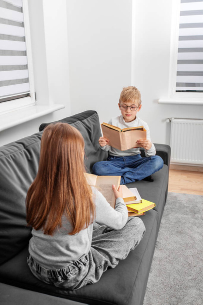 Çocuklar odadaki kanepede oturan gözlüklü kitaplar okurlar. Dünya Kitap Günü konsepti, yaşam tarzı, eğitim, eğitim. Erkek ve kız kardeş ev ödevi çalışıyor.. - Fotoğraf, Görsel