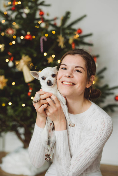 Beyaz giyinmiş, elinde küçük beyaz bir chihuahua ile Noel ağacının altında oturan gülümseyen beyaz kadın. Dikey fotoğraf. - Fotoğraf, Görsel