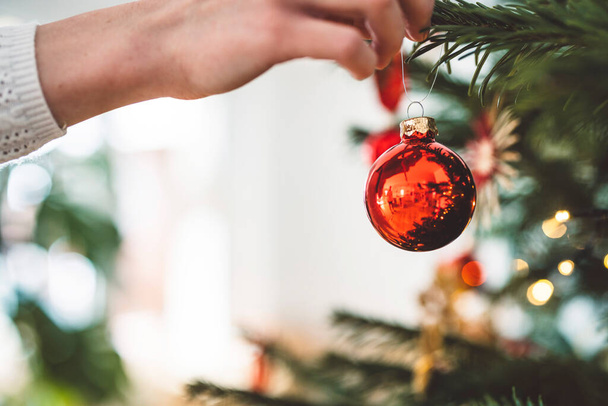 Mujer caucásica irreconocible decorando el árbol de Navidad, poniendo adorno rojo en el árbol, sosteniendo un adorno rojo de Navidad.  - Foto, imagen