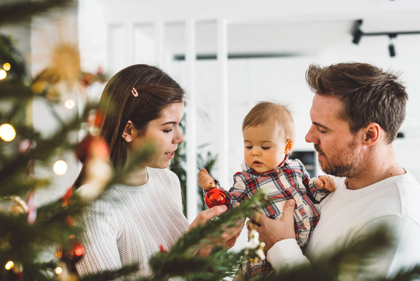 Feliz joven alegre familia caucásica de tres padres y una niña divirtiéndose decorando el árbol de Navidad. Retrato de Navidad familiar.  - Foto, Imagen