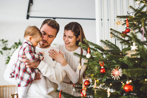 3人のお母さんのお父さんと赤ちゃんの女の子の幸せな若い陽気な白人の家族は、クリスマスツリーを飾るのを楽しんでいます。家族クリスマスの肖像画.  - 写真・画像