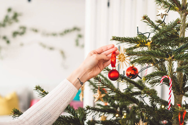 クリスマスツリーを飾る認識できない白人女性は、赤い飾りを木に置き、赤いクリスマスの飾りを保持します.  - 写真・画像