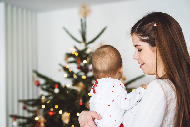 Portrait of young mom and her daughter, baby girl having fun on Christmas, decorating the Christmas tree in festive outfits. Smiling baby girl playing with her mom.  - Фото, изображение
