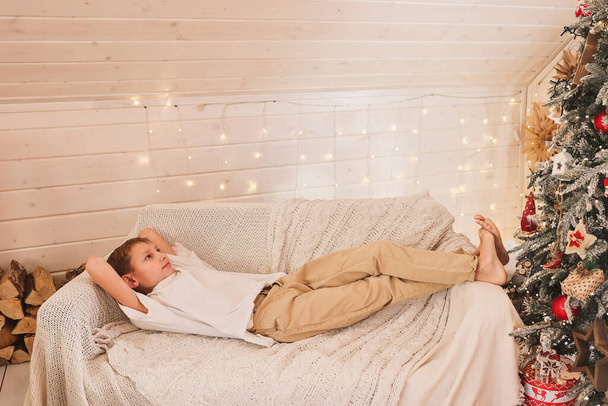 Симпатичный мальчик лежит на диване у елки в новогодней украшенной комнате - Фото, изображение