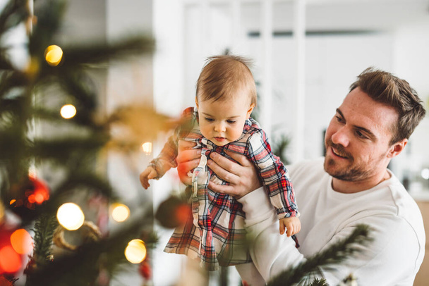 Father and daughter, baby girl having fun on Christmas, decorating the Christmass tree in festive outfits. Smiling baby girl playing with her dad.  - Photo, Image