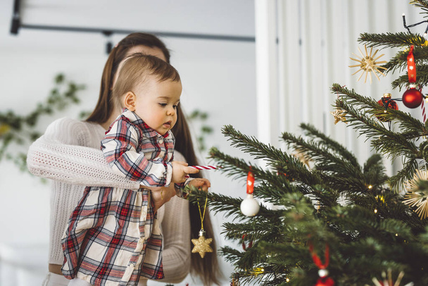 Portrait of young mom and her daughter, baby girl having fun on Christmas, decorating the Christmas tree in festive outfits. Smiling baby girl playing with her mom.  - Photo, Image