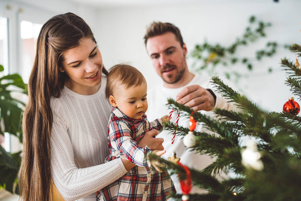 Feliz joven alegre familia caucásica de tres padres y una niña divirtiéndose decorando el árbol de Navidad. Retrato de Navidad familiar.  - Foto, Imagen