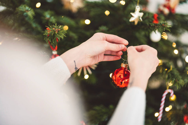 Unerkennbare kaukasische Frau schmückt den Weihnachtsbaum, setzt roten Schmuck an den Baum und hält einen roten Weihnachtsschmuck in der Hand.  - Foto, Bild