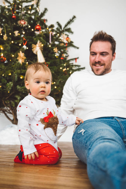 Father and daughter, baby girl having fun on Christmas, decorating the Christmass tree in festive outfits. Smiling baby girl playing with her dad.  - Photo, Image