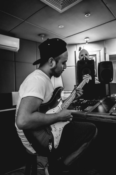 Joven músico tocando la guitarra eléctrica frente a una mesa de mezclas de sonido con varias perillas en un estudio (en blanco y negro) - Foto, imagen