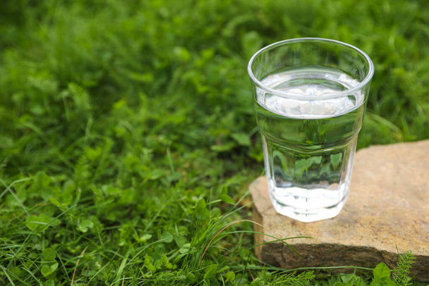 Glass of fresh water on stone in green grass outdoors. Space for text - Foto, Bild