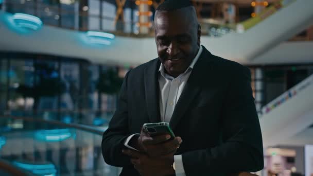 Afrikai amerikai üzletember áll bevásárlóközpont cég irodájában nézi a mobiltelefon képernyő beszélgetés videohívás okostelefon konferencia beszél barátságos mosoly válasz mobiltelefon chat - Felvétel, videó
