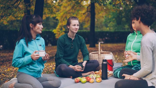 Cheerful young women friends are resting on mats having picnic talking and drinking tea after yoga class outdoors on beautiful autumn day. Food and basket are visible. - Zdjęcie, obraz
