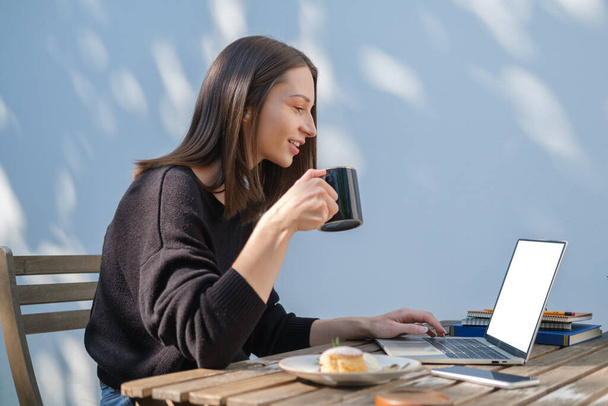 Vista lateral da jovem mulher feliz bebendo café e navegando na internet no laptop ao ar livre. - Foto, Imagem
