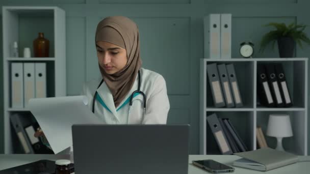 Fiatal orvos arab muszlim vallás nő csinál papírmunka a klinikán, hogy a tudományos kutatás olvasni papír dokumentumok orvosi biztosítás női orvos orvos használata laptop modern technológia az egészségügyben - Felvétel, videó