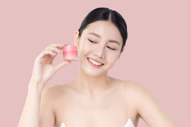 Studio shot Belle jeune femme asiatique avec une peau propre et fraîche montrant pot de crème isolé sur fond rose, Soins du visage beauté et concept de spa. - Photo, image