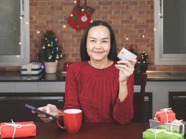 Retrato de mujer asiática con suéter rojo sentada con taza roja de café y cajas de regalo en la cocina con decoración navideña, sosteniendo teléfono móvil y tarjeta de crédito, sonriendo y mirando a la cámara. - Foto, imagen