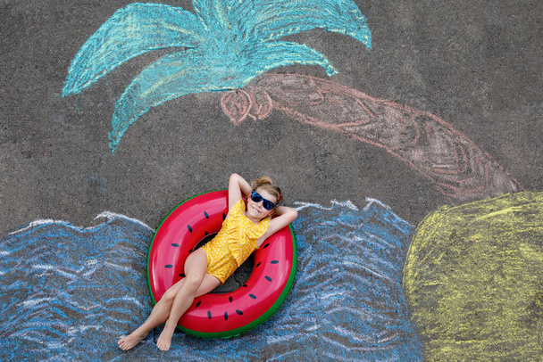Happy little preschool girl in swimsuit on inflatable ring with sea, sand, palm painted with colorful chalks on asphalt. Cute child with having fun with chalk picture. Summer, vacations, summertime. - Photo, Image