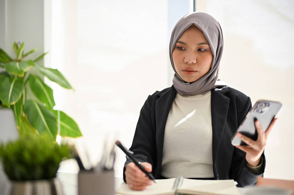 Asiatisch-muslimische Geschäftsfrau trägt Hijab, hält ihr Smartphone, macht sich Notizen auf ihrem Notizblock, konzentriert sich auf ihre Aufgaben. - Foto, Bild