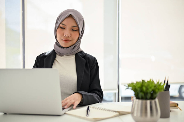 Femme d'affaires musulmane asiatique avec succès et confiante portant le hijab, utilisant un ordinateur portable, se concentrant sur ses tâches, travaillant dans le bureau. - Photo, image