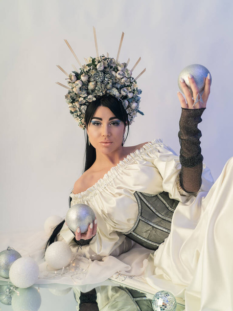 Eine schöne Brünette im Bild einer Schneekönigin mit Krone und einem weißen Seidenkleid mit Korsett. Die Schönheit des Gesichts. Wintermärchen. Ein fabelhaftes Bild.  - Foto, Bild