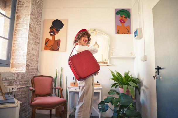 Young, beautiful, bohemian Latina, wearing a red beret on her head and a red vintage suitcase, looking happily at the camera for the photo. Concept of an enterprising woman. - Foto, Imagem
