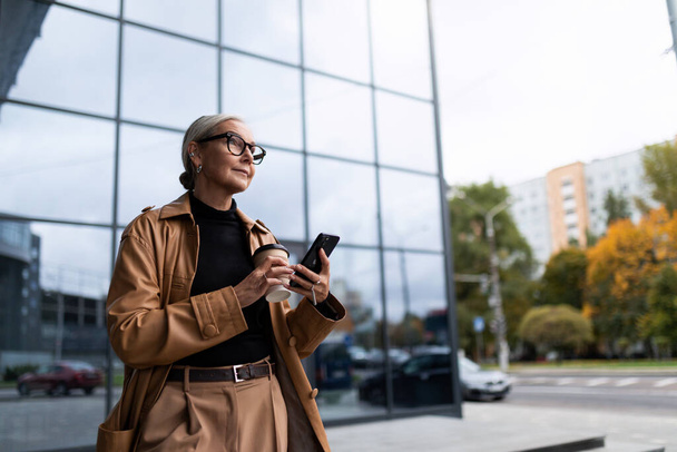 elegante mujer de negocios senior caucásica en un abrigo de cuero marrón con un teléfono móvil en el fondo del centro de oficina exterior. - Foto, imagen