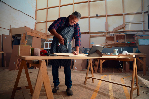 Multiracial hombre adulto medio sonriendo mientras se utiliza taladro en madera junto a la tableta digital en la mesa en la fábrica de carpintería - Foto, imagen