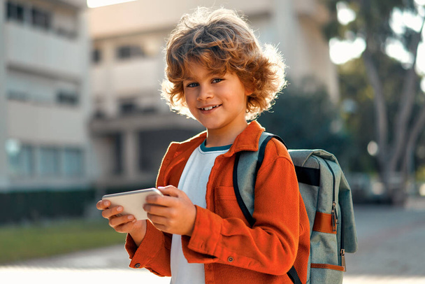 Back to school. Cute kid with stylish hairstyle with backpack holding mobile phone playing with mobile phone outdoors. Schoolboy pupil with a bag. Elementary school student after class. - 写真・画像