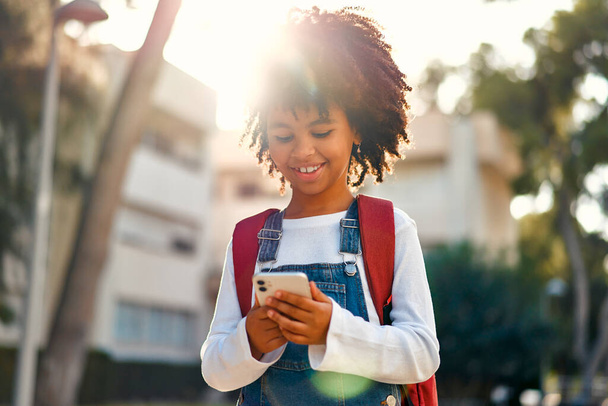 Vissza a suliba. Boldog aranyos afro-amerikai okos lány hátizsák tartja mobiltelefon játszik mobiltelefon a szabadban. Iskoláslány egy táskával. Általános iskolai hallgató óra után. - Fotó, kép