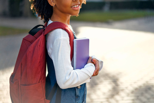 Happy cute African American smart girl with a curly afro hairstyle, a school bag and notebooks in her hands on the street. Modern backpack. First time to school. Back to school. - Фото, изображение