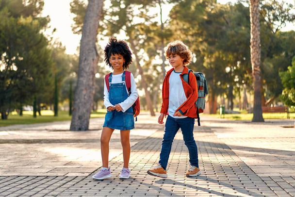 Boldog okos gyerekek iskolatáskákkal rohannak az iskolába. Vissza a suliba. Egy afro-amerikai iskolás lány az osztálytársával sétál az utcán suli után.. - Fotó, kép