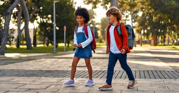 Χαρούμενα έξυπνα παιδιά με σχολικές τσάντες σπεύδουν στα μαθήματα στο σχολείο. Πίσω στο σχολείο. Μια Αφρο-Αμερικανίδα μαθήτρια με τον συμμαθητή της περπατούν στο δρόμο μετά το σχολείο.. - Φωτογραφία, εικόνα