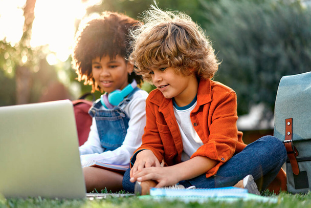 Urocze uczennice razem odrabiają lekcje używając laptopa, siedząc na trawie w parku. Wracać do szkoły. Afroamerykanka dziewczyna z kaukaski słodkie chłopiec komunikować się po szkole. - Zdjęcie, obraz