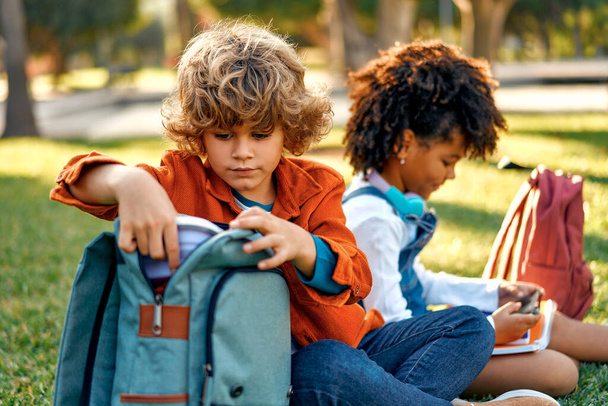 Charming schoolchildren after lessons are resting sitting on the grass in the park. Back to school. African American girl with a Caucasian boy take out a lunch box from their backpack to have lunch. - Φωτογραφία, εικόνα
