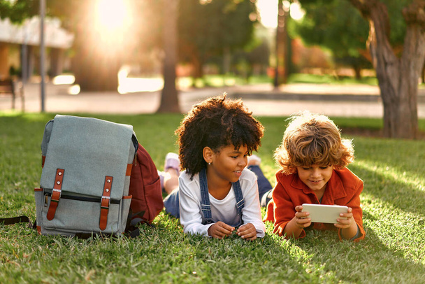 Adorable schoolchildren play games on the phone while lying on the grass in the park. Back to school. African American girl with Caucasian cute boy having fun after school. - Φωτογραφία, εικόνα