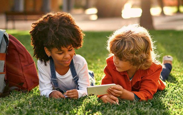 Schattige schoolkinderen spelen spelletjes aan de telefoon terwijl ze op het gras in het park liggen. Terug naar school. Afro-Amerikaans meisje met Kaukasisch schattig jongen hebben plezier na school. - Foto, afbeelding