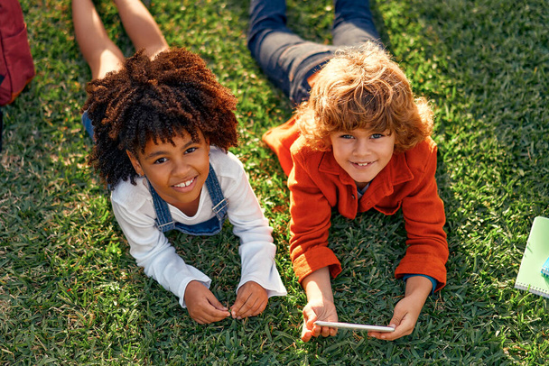 Suloiset koululaiset pelaavat pelejä puhelimessa makatessaan puistossa nurmella. Takaisin kouluun. Afrikkalainen amerikkalainen tyttö valkoihoinen söpö poika hauskaa koulun jälkeen. - Valokuva, kuva