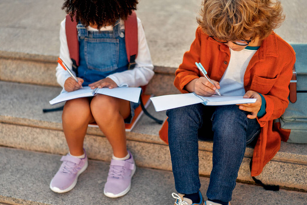 Charming smart schoolchildren doing their homework together while sitting on the stairs near the school. Back to school. African American girl with caucasian boy in glasses studying together. - Foto, imagen