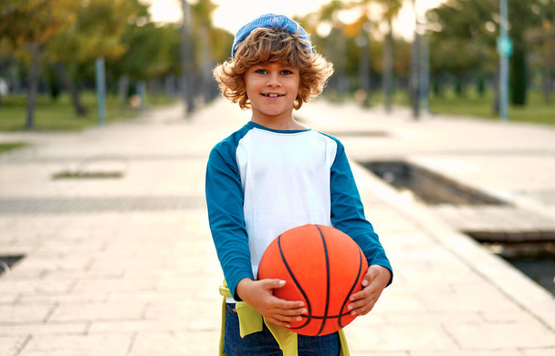 スポーツとバスケットボール。公園の屋外にバスケットボールボールを持つスタイリッシュなかわいい男の子立っている。スポーツ、運動、健康的なライフスタイル、広告、アクション、運動の概念. - 写真・画像