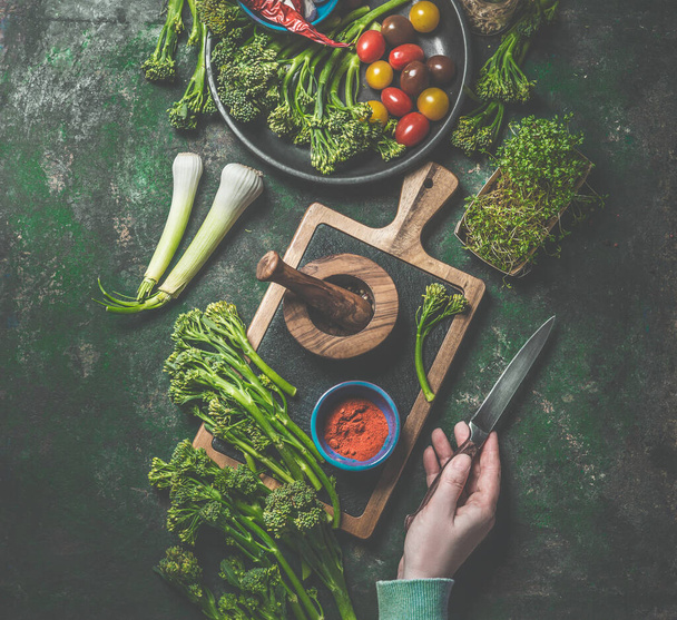Cooking preparation of wild broccoli. Women hand holding knife on dark rustic background with cutting board and ingredients. Top view. Healthy food - Foto, afbeelding