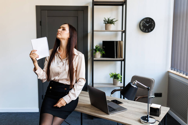 Mid adult businesswoman leaning on desk fanning papers in office showing that office is very hot. - Photo, image