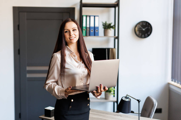 Adult successful female employee standing in modern office, hold a laptop in arms. Happy brunette-hair business lady in elegant stylish suit looks at camera and smiling - Foto, Imagem
