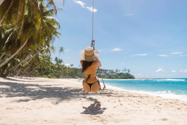 Bikini beach girl model in black swimsuit having fun on swing on palm tree bungee on tropical sea shore during summer vacations. High quality banner photo with copy space, clear sky for text.  - Fotoğraf, Görsel