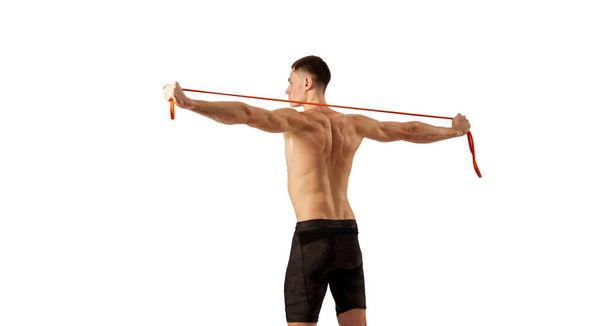 Back view of muscular young man doing excersice with expander rope isolated over white background. Fitness, healthy and active lifestyle concept. Shirtless athlete wearing bicycle shorts - Photo, Image