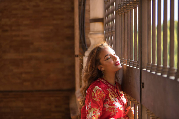 Young and beautiful woman in a typical Moroccan red suit, with embroidery with gold and silver threads, leaning on a grille receiving the sun's rays. Concept ethnicity, typical costumes, Arabic. - Foto, afbeelding