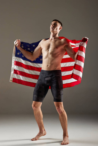 Retrato del joven deportista de pie con la bandera de Estados Unidos sobre sus hombros y expresando las emociones ganadoras aisladas sobre un fondo gris. Concepto de victoria, victoria, éxito, deporte, logros - Foto, Imagen