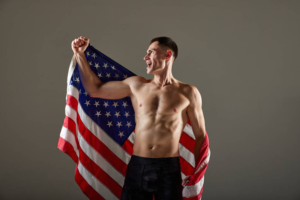 Портрет молодого спортсмена, стоящего с флагом США на плечах и выражающего эмоции победителя на сером фоне. Концепция победы, победы, успеха, спорта, достижений - Фото, изображение