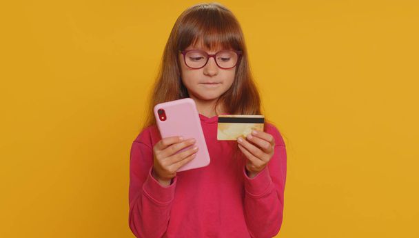 Young preteen child girl kid using credit bank card and smartphone while transferring money, purchases online shopping, order food delivery. Little toddler children alone on studio yellow background - Fotografie, Obrázek