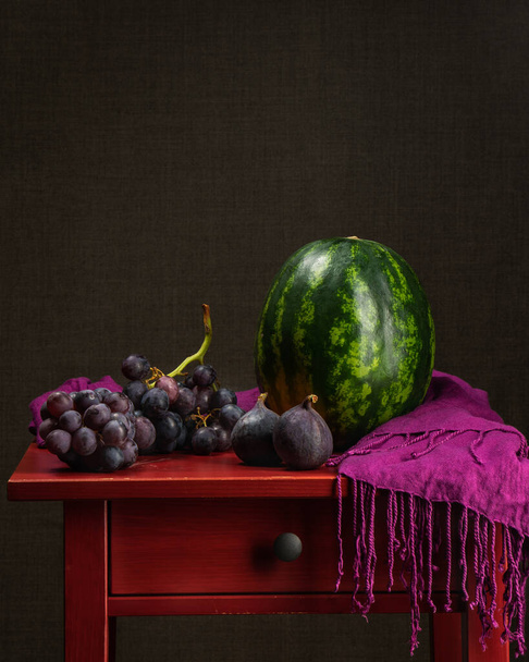 Duży owalny arbuz, grono winogron. Dwie dojrzałe figi na czerwonym drewnianym stole. Obok jest ciemnoróżowy szal. Właściwe odżywianie. Jedzenie. Wegańskie. - Tak. Zdrowe jedzenie. Witaminy. Zdrowie. - Zdjęcie, obraz